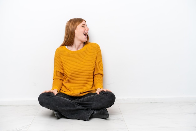 Young redhead woman sitting on the floor isolated on white background laughing in lateral position