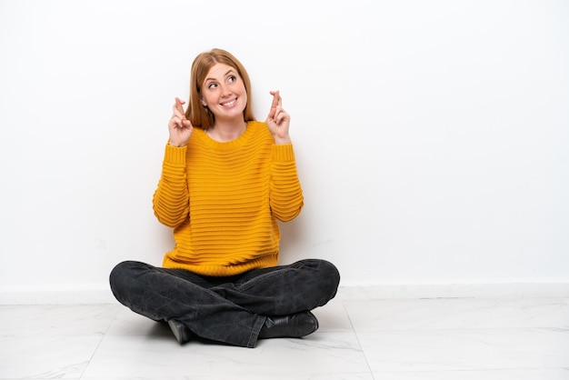 Young redhead woman sitting on the floor isolated on white background with fingers crossing