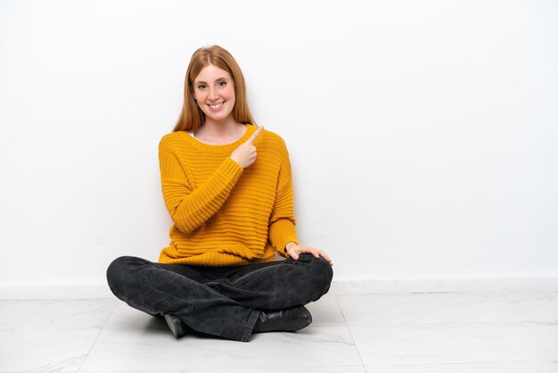 Фото Молодая рыжая женщина сидит на полу на белом фоне, указывая в сторону, чтобы представить продукт