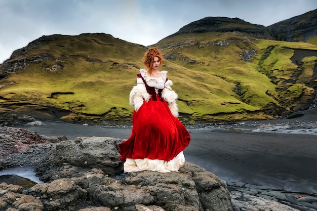 A young redhead woman stays on big stone near ocean coastline in old-fashioned clothes. Faroe Islands, Denmark