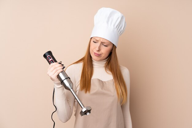 Young redhead woman using hand blender with sad expression