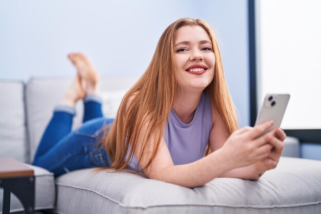 Молодая рыжая женщина с смартфоном лежит на диване дома