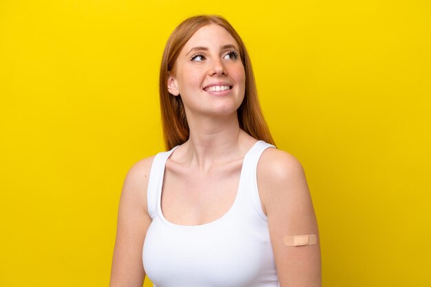 Young redhead woman wearing a bandaids isolated on yellow background looking up while smiling