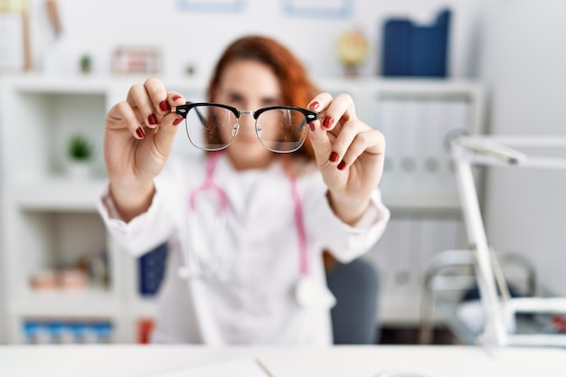 Young redhead woman wearing doctor uniform holding glasses at hospital