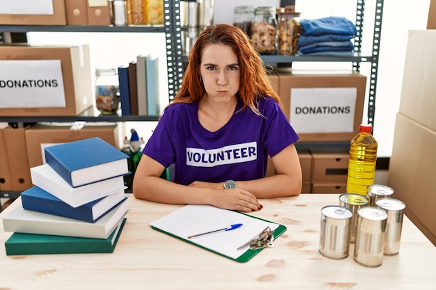 Photo young redhead woman wearing volunteer t shirt at donations stand puffing cheeks with funny face. mouth inflated with air, crazy expression.