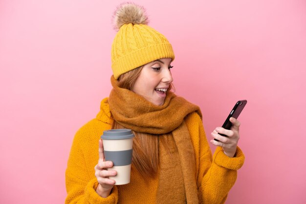 Giovane donna rossa che indossa una giacca invernale isolata su sfondo rosa che tiene il caffè da portare via e un cellulare