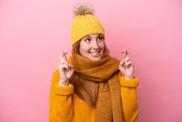 Young redhead woman wearing winter jacket isolated on pink background with fingers crossing