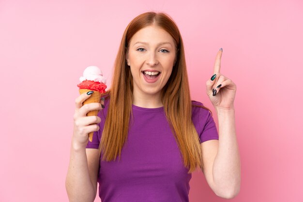 좋은 아이디어를 가리키는 격리 된 분홍색 벽 위에 코 넷 아이스크림을 가진 젊은 빨강 머리 여자