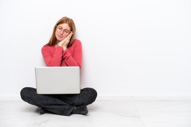 Young redhead woman with a laptop sitting on the floor isolated on white background making sleep gesture in dorable expression