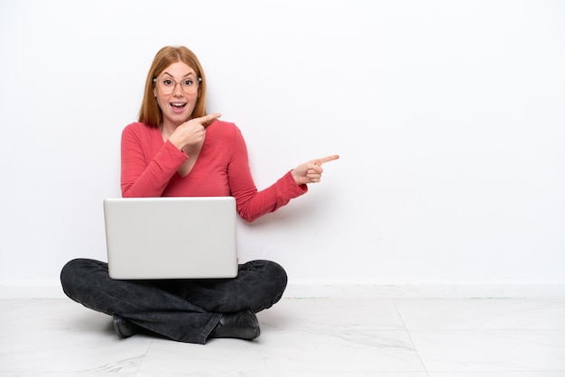 Young redhead woman with a laptop sitting on the floor isolated\
on white background surprised and pointing side