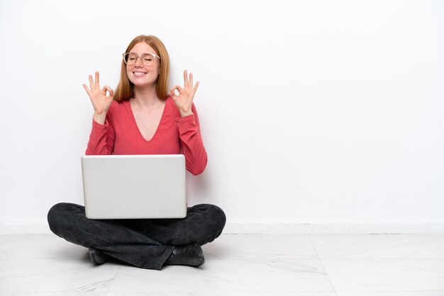 Young redhead woman with a laptop sitting on the floor isolated on white background in zen pose