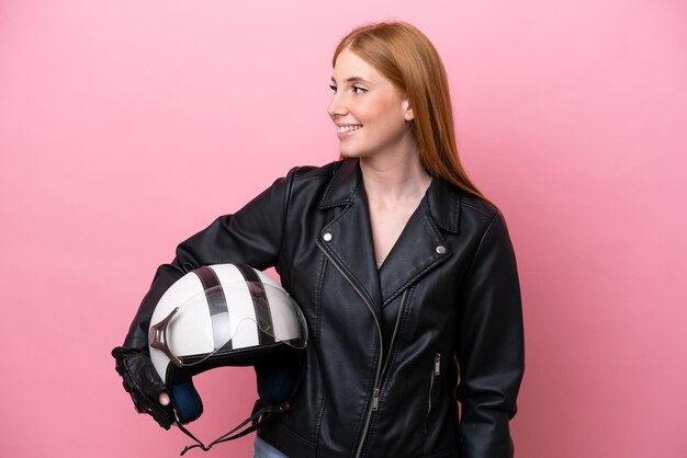 側を見ているピンクの背景に分離されたオートバイのヘルメットを持つ若い赤毛の女性