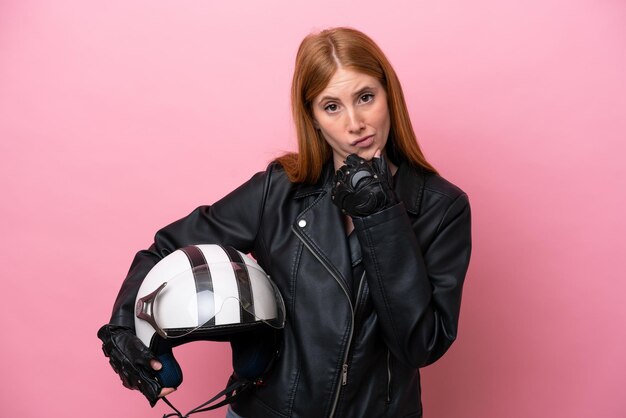 Young redhead woman with a motorcycle helmet isolated on pink\
background thinking