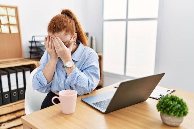 울면서 손으로 얼굴을 가리는 슬픈 표정으로 컴퓨터 노트북을 사용하여 사무실에서 일하는 젊은 빨간머리 여성. 우울증 개념.