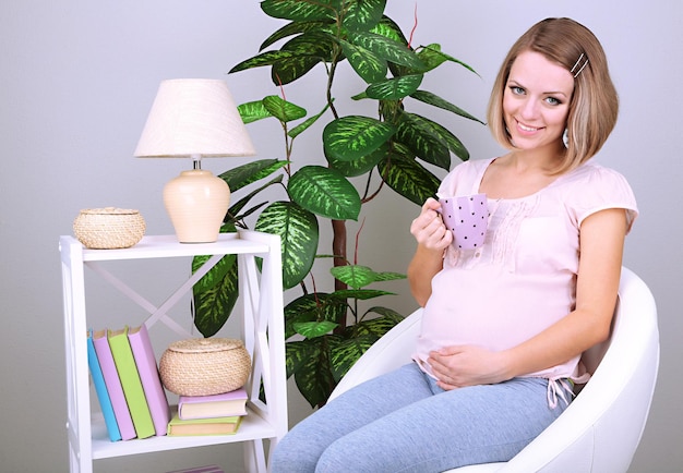 Young regnant woman sitting on armchair and holding cup at home