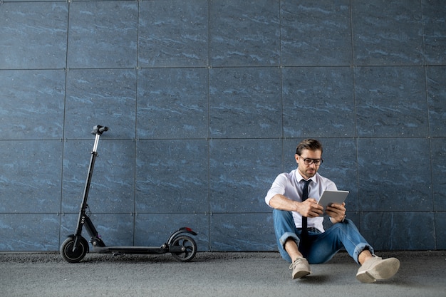 Молодой расслабленный бизнесмен в элегантной повседневной одежде сидит у стены здания и прокручивает в цифровом планшете