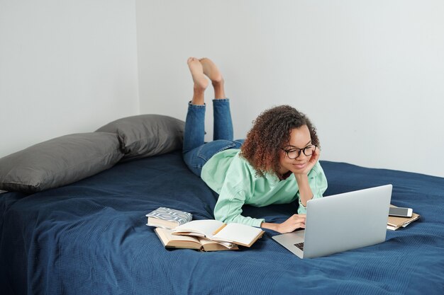 ノートパソコンの前のベッドに横たわって、次の試験の準備をしながらネットサーフィンをしているジーンズとパーカーの若いリラックスした女子学生