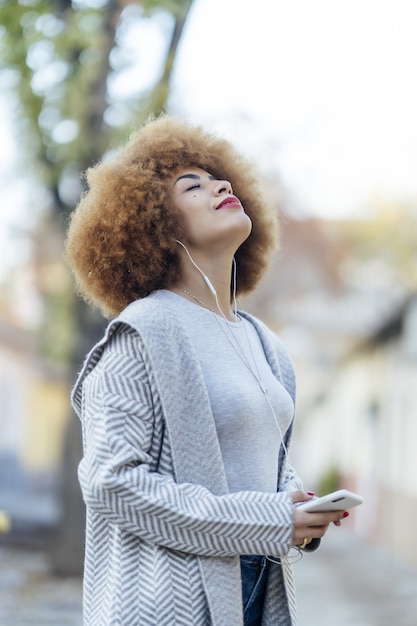 Young relaxed Hispanic female with curly hair listening to music with headphones in a park