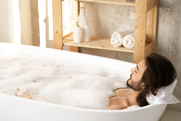 Young relaxed man enjoying bath with foam while putting his head on soft rolled white towel