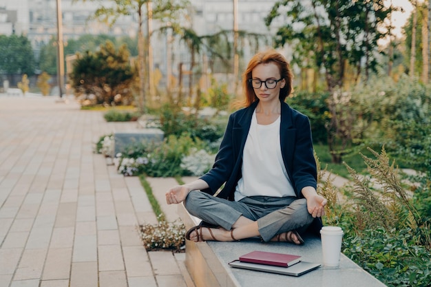 Young relaxed redhead woman meditates in park feels calm sits in lotus pose outdoor