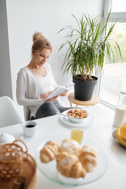 朝、自宅で健康的な朝食を食べた後、台所のテーブルのそばに座って読んでリラックスした若い女性