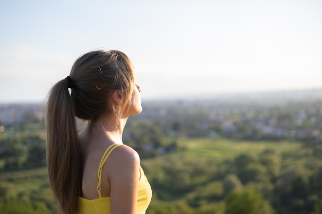 緑の野原に立つ若いリラックスした女性が、夜の自然の中で夕日を眺める。