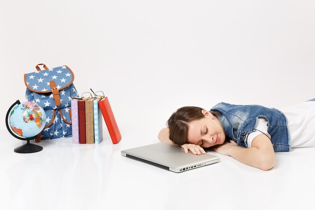 Молодая расслабленная студентка в джинсовой одежде, спящая на портативном компьютере, лежащем рядом с земным шаром, рюкзаком, школьными учебниками