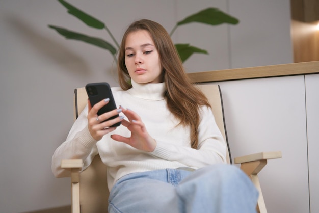 Young relaxed woman teenager using smartphone cell mobile phone at home in comfortable armchair