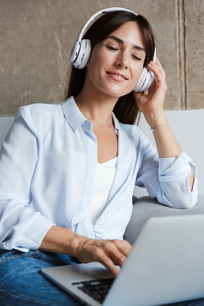 young relaxing woman indoors at home sit on sofa in living room listening music with headphones using laptop computer.