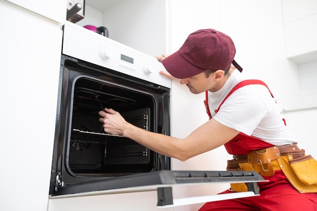 Photo young repairman in protective workwear fixing oven in kitchen.