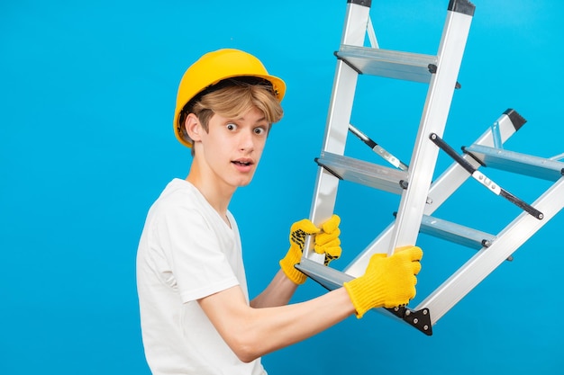 白い t シャツと青い背景にスタジオに立っている手にはしごを保持している頭に黄色のヘルメットと手袋の若い修理工 10 代青の背景に分離された将来の建築家