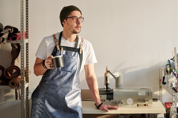 Giovane artigiano riposante in grembiule in pelle e cappello beanie che tiene tazza con bevanda mentre è in piedi al tavolo in officina