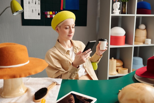 Young restful craftswoman using smartphone and having coffee