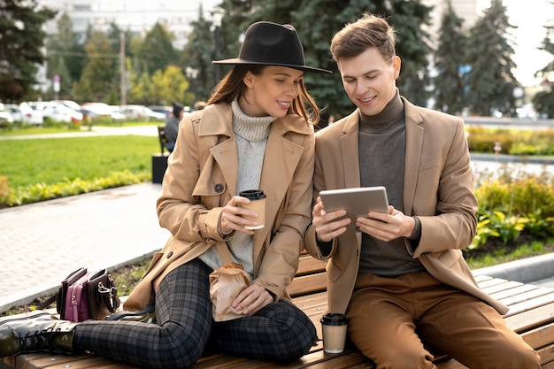 Young restful female looking at screen of smartphone held by her boyfriend while both sitting on bench and having chat and coffee