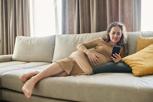 Young restful pregnant woman in casual dress relaxing on soft couch