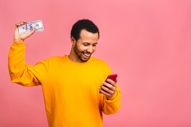 Молодой богатый человек в повседневной одежде держит долларовые банкноты и мобильный телефон с удивлением, изолированным над розовой стеной