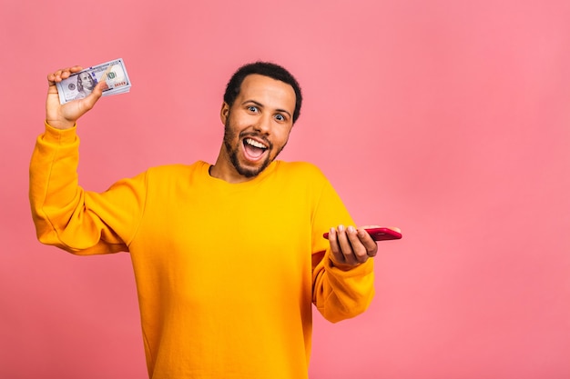 Young rich man in casual holding money dollar bills and mobile phone with surprise isolated over pink wall