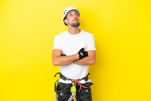 Young rock- climber man isolated on yellow background looking up while smiling