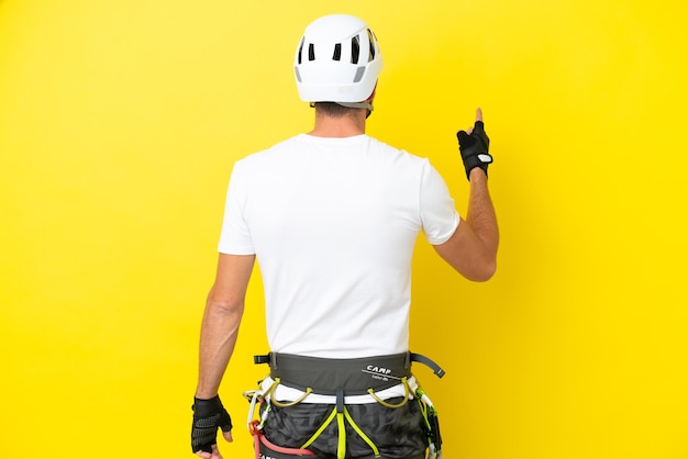 Young rock- climber man isolated on yellow background pointing\
back with the index finger