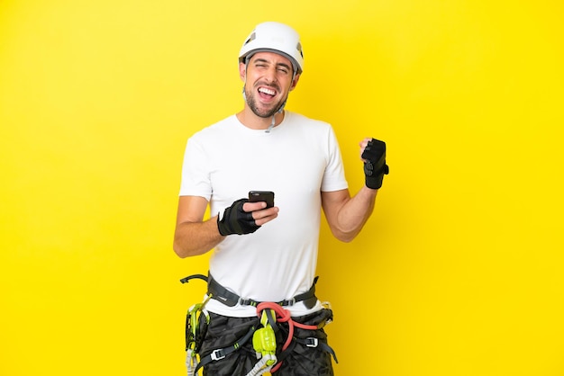 Young rock- climber man isolated on yellow background with phone in victory position