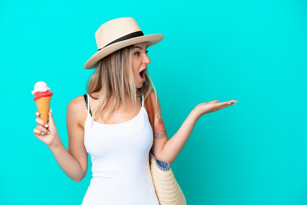 Young Romanian woman holding ice cream and beach bag isolated on blue background with surprise facial expression