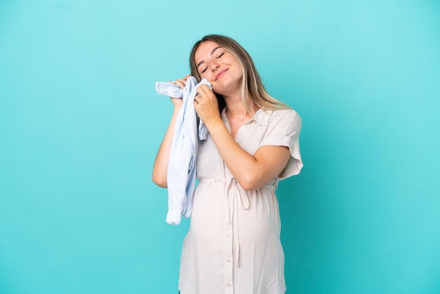 Young Romanian woman isolated on blue background pregnant and holding baby clothes