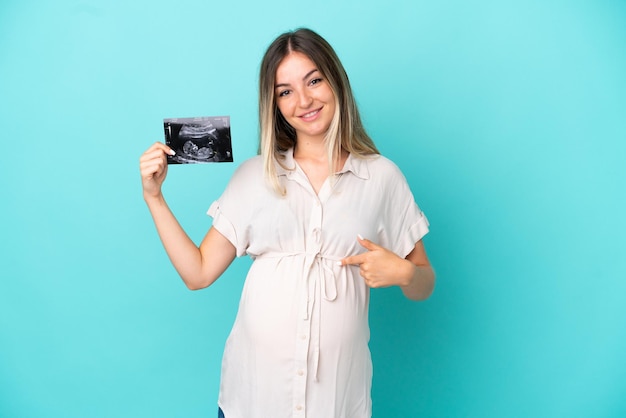 Young Romanian woman isolated on blue background pregnant and holding an ultrasound