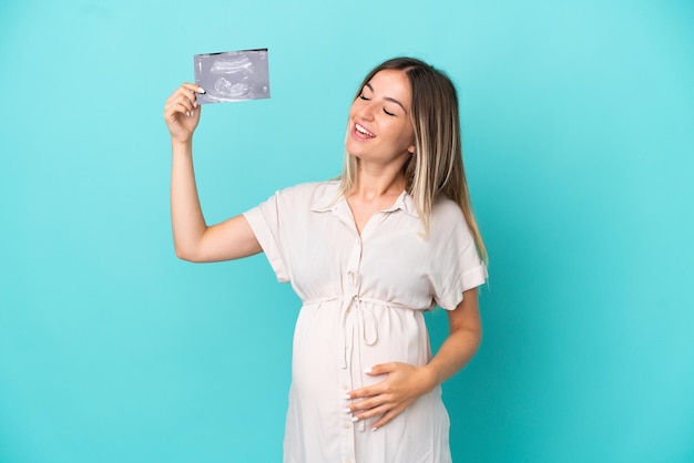 Young Romanian woman isolated on blue background pregnant and holding an ultrasound