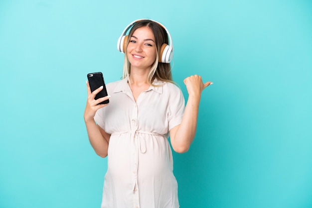 Young Romanian woman isolated on blue background pregnant and listening music while pointing side