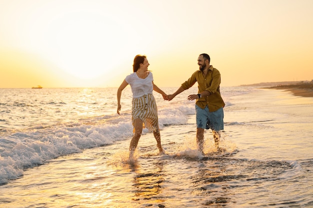 海のそばを振り返って踊る若いロマンチックなカップル美しい空と夕日の海の景色黄金の夕日のビーチでロマンチックなカップルビーチで楽しんでいる恋人のカップル