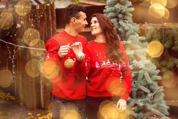 Young romantic couple is decorating Christmas tree outdoors before Christmas. They holding balls on the tree and smiling.