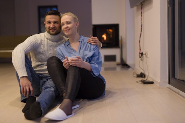 Young romantic couple sitting on the floor in front of fireplace,talking and drinking tea on cold winter night at home