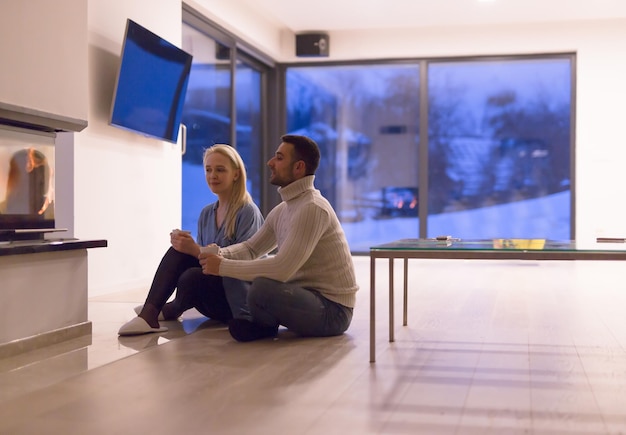 Young romantic couple sitting on the floor in front of fireplace,talking and drinking tea on cold winter night at home