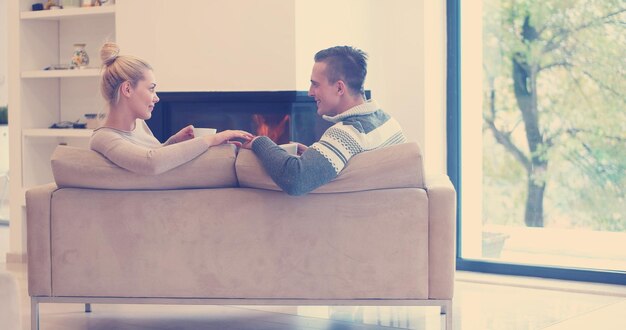 Young romantic couple sitting on sofa in front of fireplace at\
home, looking at each other, talking and drinking coffee at autumn\
day